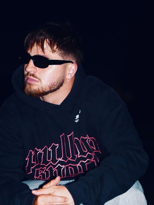 Man wearing sunglasses and a black hoodie with red text sits on a road at night, ambient lights in the background, looking contemplative.