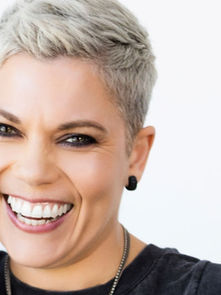 Smiling person with short blonde hair, wearing black earrings and a bracelet, against a white background. Bright and cheerful mood.