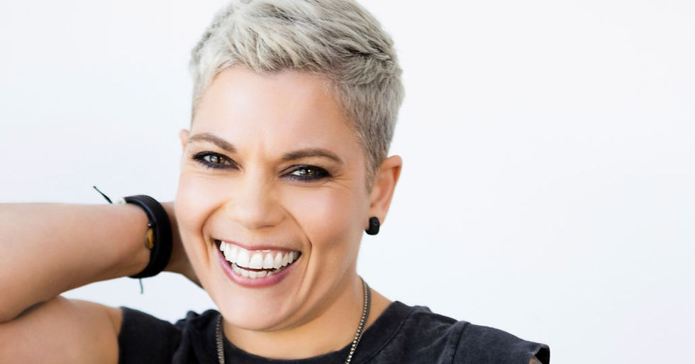 Smiling person with short blonde hair, wearing black earrings and a bracelet, against a white background. Bright and cheerful mood.