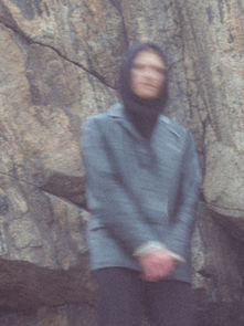 Blurred person in a coat and headscarf stands in front of a textured rock wall. The image conveys a mysterious, serene mood.