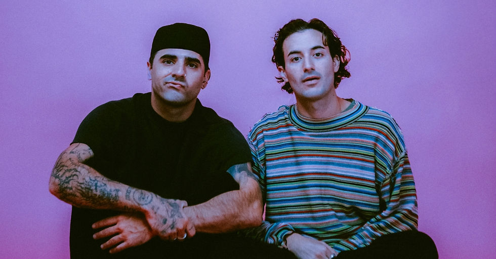 Two men sit against a pink background. One wears a black shirt and beanie, showing tattoos; the other wears a colorful striped shirt, both serious.