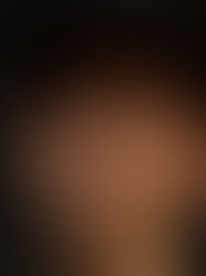 Young person with a neutral expression, wearing a dark cap and black hoodie, standing against a dark background. Face is softly lit.