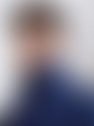 Young man in a blue jacket with a high collar stands against a light, abstract background, looking confident and thoughtful.