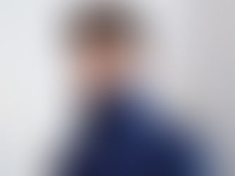 Young man in a blue jacket with a high collar stands against a light, abstract background, looking confident and thoughtful.