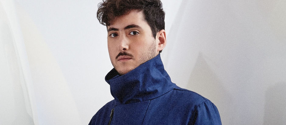 Young man in a blue jacket with a high collar stands against a light, abstract background, looking confident and thoughtful.