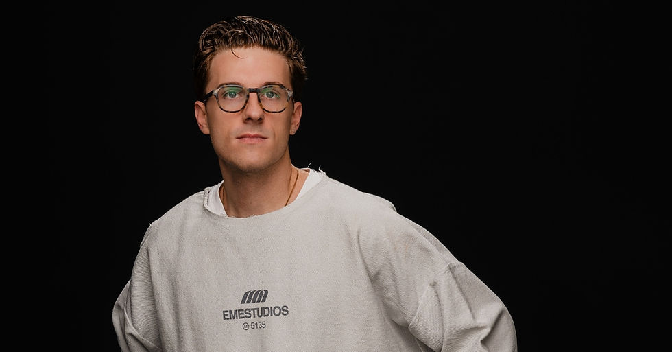Man with glasses in a light gray Emestudios sweatshirt stands against a black background, looking directly ahead. Calm expression.