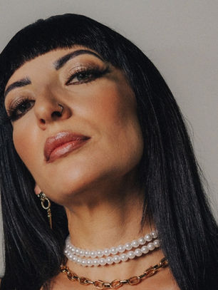 Woman with black hair gazes confidently, wearing gold and pearl necklaces, against a neutral background. Elegant and poised.