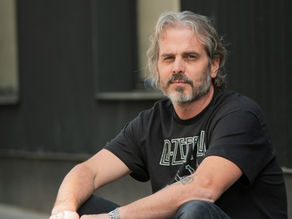 Gray-haired man with beard in a black Led Zeppelin T-shirt sits outdoors against a dark wall, looking calm.