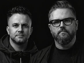 Black-and-white portrait of two serious men against a dark background, one wearing glasses and a jacket.