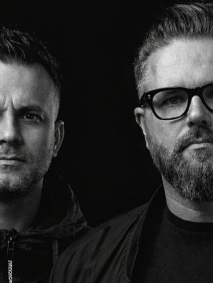 Black-and-white portrait of two serious men against a dark background, one wearing glasses and a jacket.