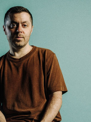 Man in brown shirt sits calmly against a teal background, conveying a relaxed mood. No visible text or patterns.