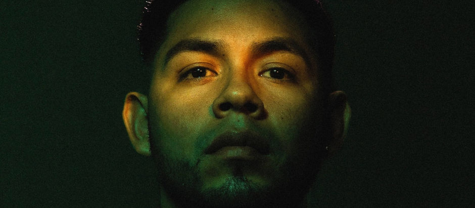 Close-up of a man's face lit with green and yellow hues against a dark background, creating a dramatic and contemplative mood.