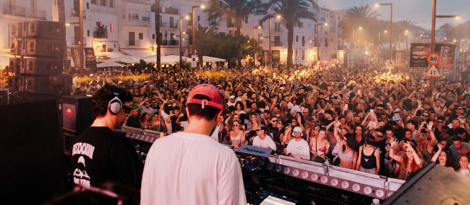 Two DJs perform for a huge cheering crowd at a smoky street party lined with palm trees and white buildings; 20 speed sign visible.
