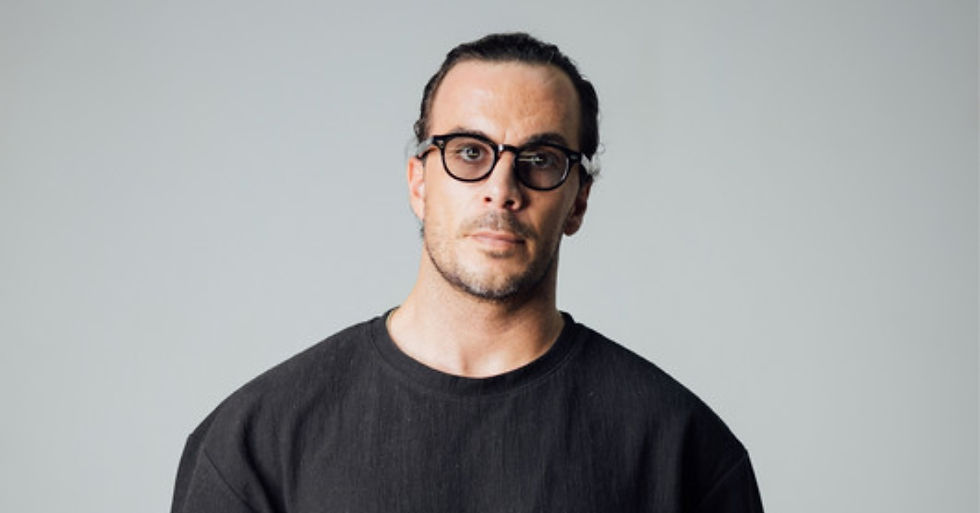 Man with glasses and tied-back hair in a black sweater against a plain gray background, looking serious.