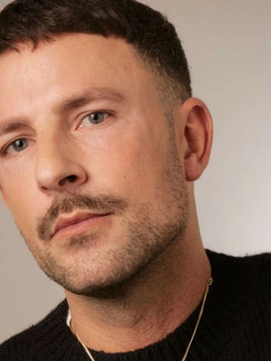 Close-up portrait of a man with short dark hair and a mustache, wearing a black sweater against a plain beige background.