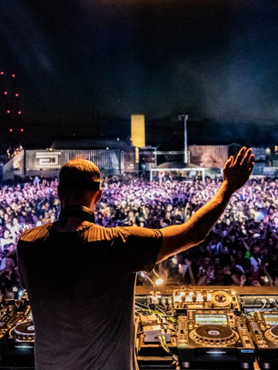 DJ with raised arm performs at a vibrant outdoor concert at night, with a huge crowd, blue lights, and music equipment in the foreground.