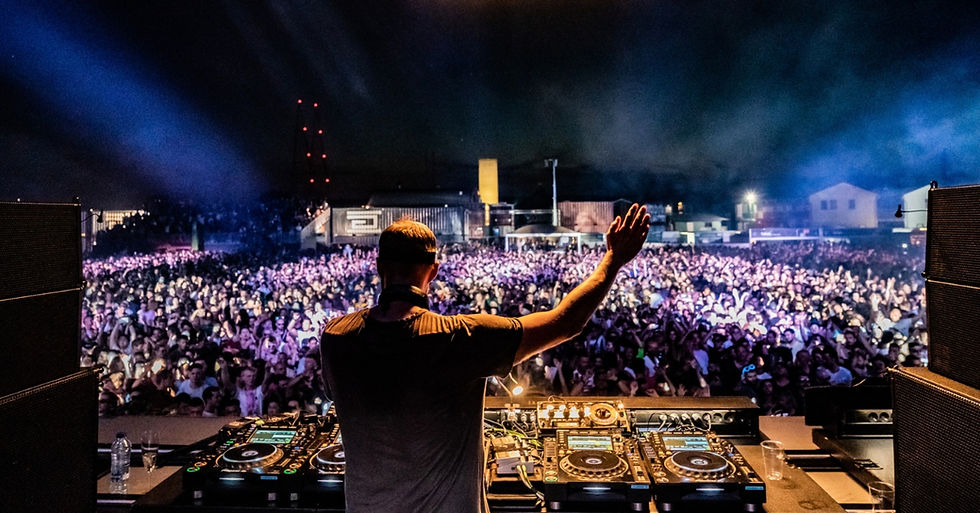 DJ with raised arm performs at a vibrant outdoor concert at night, with a huge crowd, blue lights, and music equipment in the foreground.
