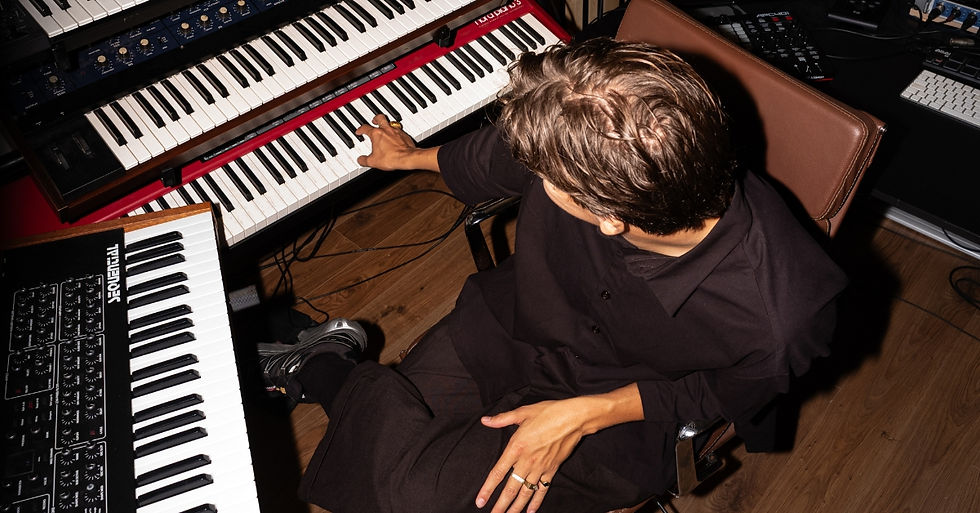 Person in black outfit playing a red and black keyboard in a studio. Multiple keyboards and equipment around, wooden floor visible.