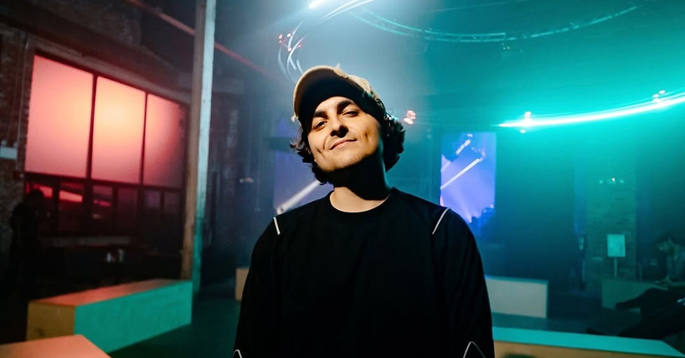 Person in a black shirt and cap smiling in a dimly lit room with colorful neon lights and a glowing screen background. Vibrant atmosphere.