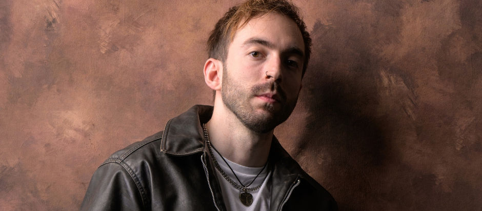 Portrait of a young man in a black leather jacket and necklaces against a textured brown backdrop, looking calmly at camera.
