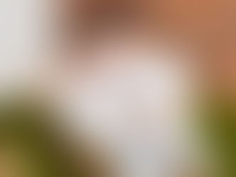 Young man in white shirt, beige pants, sitting relaxed on green couch. Brick wall background. Tattoos visible, thoughtful expression.