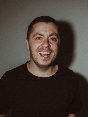 Smiling man in a black shirt against a gray background. Soft lighting creates a shadow, giving a cheerful and relaxed mood.