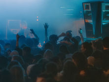Crowd dancing at a blue-lit nightclub concert, hands raised in haze beside tall speakers.