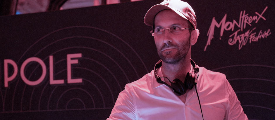Man wearing glasses and a cap, with headphones around his neck, standing in front of Montreux Jazz Festival backdrop, bathed in pink light.
