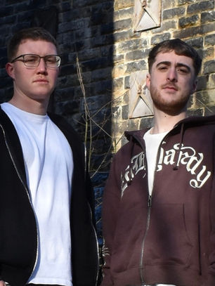 Two men stand in front of a stone wall, one wears glasses and a black hoodie, the other a brown hoodie with white lettering. Both look serious.