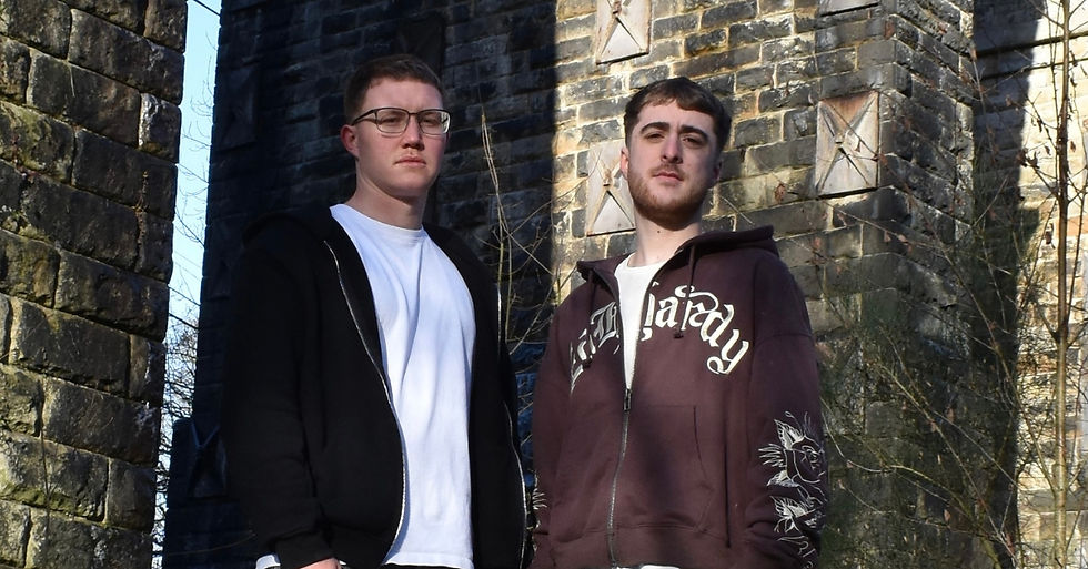 Two men stand in front of a stone wall, one wears glasses and a black hoodie, the other a brown hoodie with white lettering. Both look serious.
