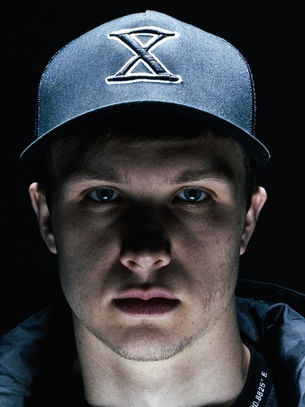 Serious young man in a dark cap with an X logo, lit against a black background, staring directly at the camera.