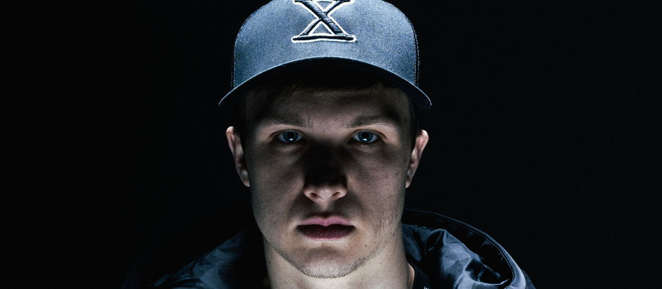 Serious young man in a dark cap with an X logo, lit against a black background, staring directly at the camera.