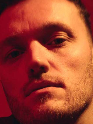 Close-up of a man's face with a serious expression, reflected in a red-tinted mirror. Warm lighting and a soft focus create an introspective mood.