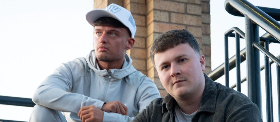 Two men sit on stairs outdoors, one in a white cap and hoodie, looking thoughtful. Brick wall and railings in the background.