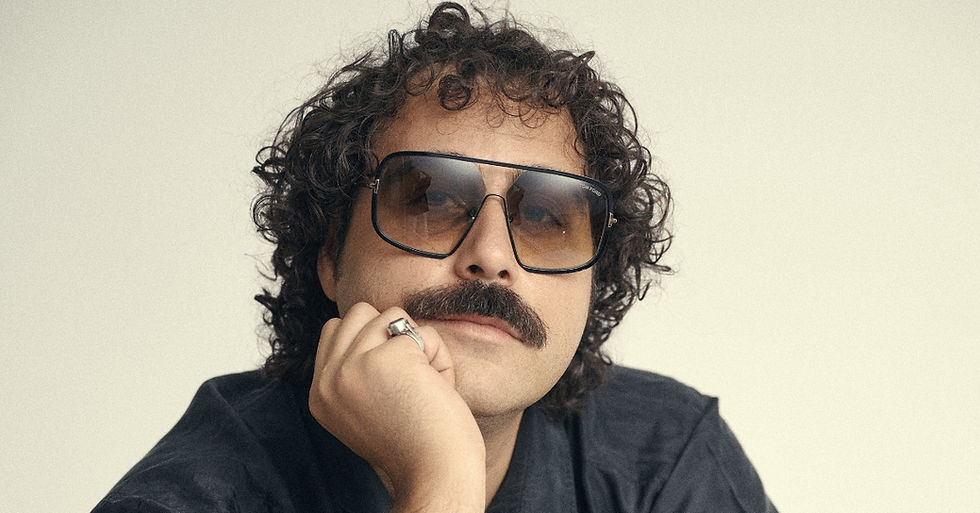 Curly-haired man with mustache and oversized sunglasses rests his chin on his hand against a plain light background.
