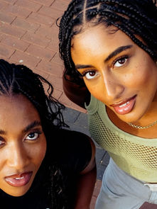 Two women with braided hair smile up at the camera on a brick pavement. One wears a green mesh top, the other a black tee. Bright, lively mood.