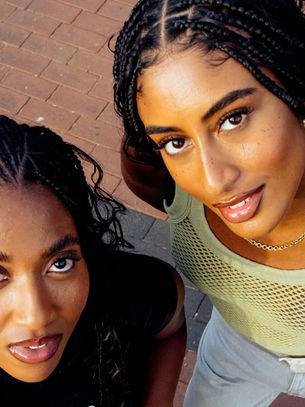 Two women with braided hair smile up at the camera on a brick pavement. One wears a green mesh top, the other a black tee. Bright, lively mood.
