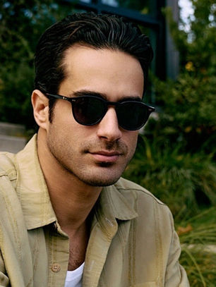 Man in sunglasses and beige shirt sits outdoors by stone steps and greenery, looking calm and composed.