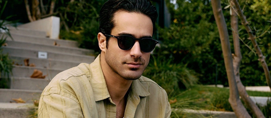 Man in sunglasses and beige shirt sits outdoors by stone steps and greenery, looking calm and composed.