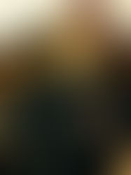 Young man with blond hair sits in a leather chair, wearing a printed black shirt. Shadowy background with plants. Calm expression.