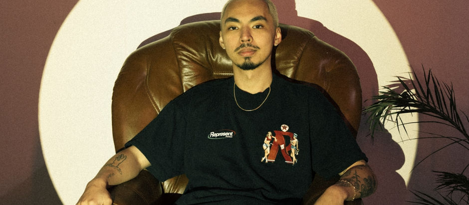 Young man with blond hair sits in a leather chair, wearing a printed black shirt. Shadowy background with plants. Calm expression.