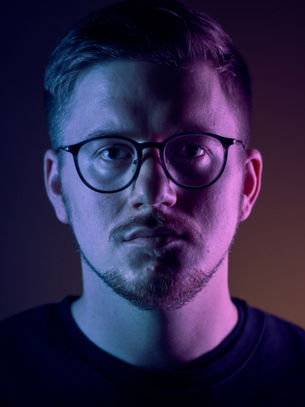 Man with glasses in purple and blue lighting, serious expression, dark background, wearing a black shirt.