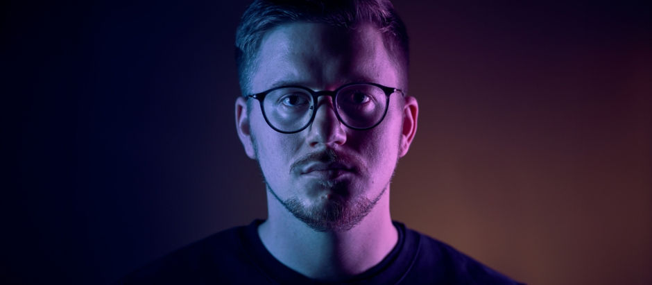 Man with glasses in purple and blue lighting, serious expression, dark background, wearing a black shirt.