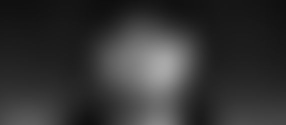 Bearded man in a black cap and hoodie stares intently at the camera against a blurred dark background. The mood is serious and contemplative.