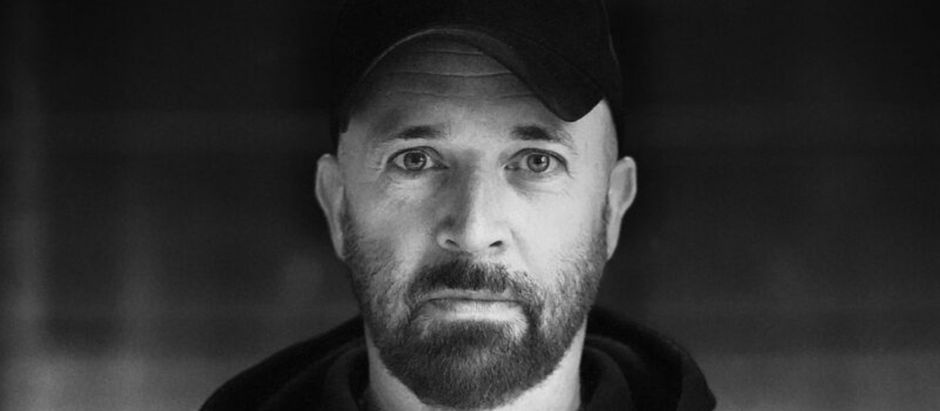 Bearded man in a black cap and hoodie stares intently at the camera against a blurred dark background. The mood is serious and contemplative.