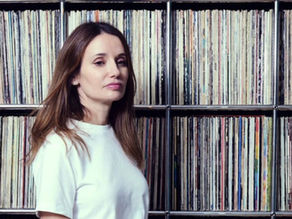 Woman in a white shirt stands before shelves packed with vinyl records, looking calm in a music-filled room.