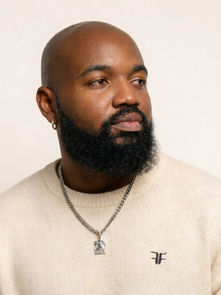 Bald man with a beard wearing a cream sweater and a necklace against a plain background. He is looking to the side, expression thoughtful.