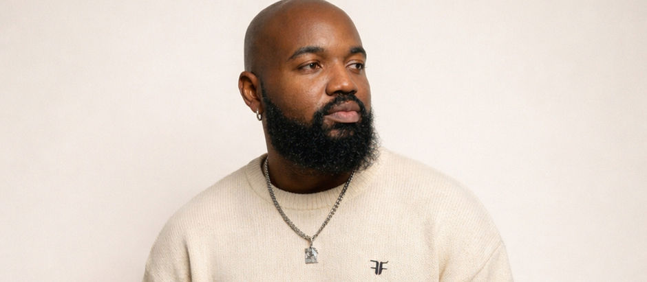 Bald man with a beard wearing a cream sweater and a necklace against a plain background. He is looking to the side, expression thoughtful.