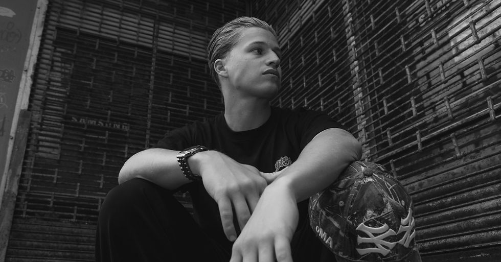 Young man in a black shirt sits against a metal gate, gazing sideways. He wears a watch and holds a patterned cap. Monochrome tone.