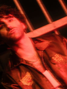 Man in a red-lit setting, leaning against metal bars, wearing a jacket with embroidered patterns. Moody and blurred background.